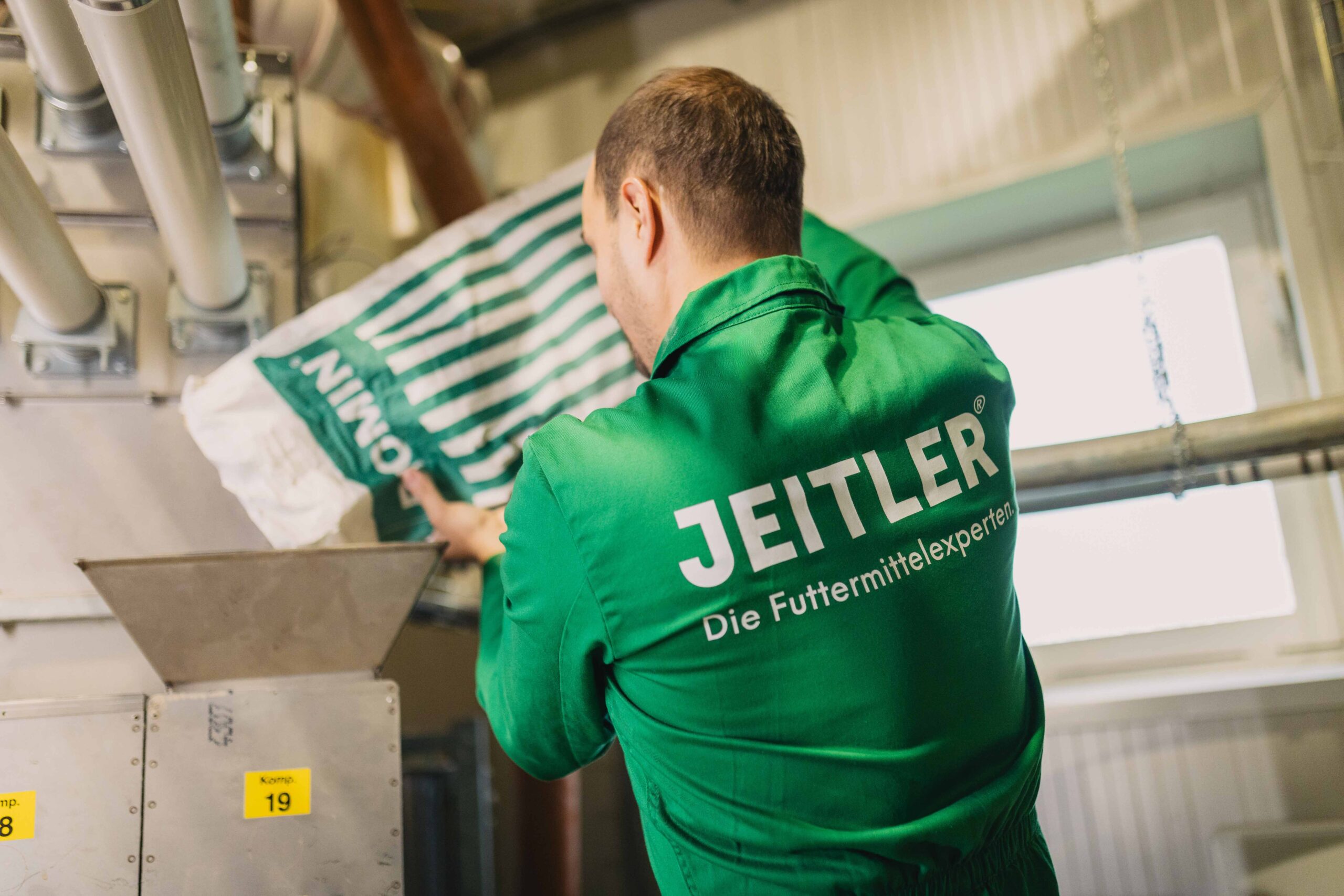 Ein Mitarbeiter in grüner Arbeitskleidung mit dem Logo "JEITLER – Die Futtermittelexperten" leert einen grünen Futtersack in einen Trichter